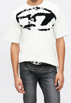 Homme portant un t-shirt blanc oversize avec un imprimé noir abstrait, un jean foncé délavé et une ceinture noire avec une boucle circulaire blanche.