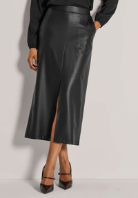 Schwarzer Leder-Midirock mit seitlichem Schlitz, glatter Textur und enganliegendem Taillenbund. Kombiniert mit schwarzen spitzen High Heels.
