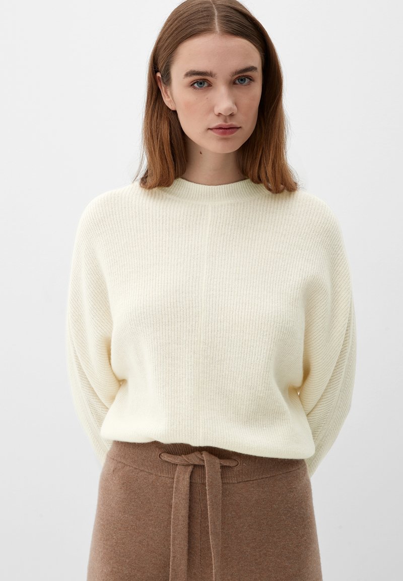 Cremefarvet strikket trøje med ribbede teksturer, rund halsudskæring og afslappet pasform, parret med en brun uldskørt med bånd i taljen.