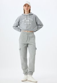 Grijze cropped hoodie met de opdruk "New York 86 RUN THE BRONX" en grijze joggers met zijzakken en elastische boorden. Witte sneakers.