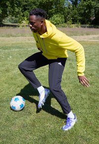 Atleta in un luminoso hoodie giallo e pantaloni da ginnastica neri, che palleggia una pallone da calcio con scarpini viola e argentati su erba verde.
