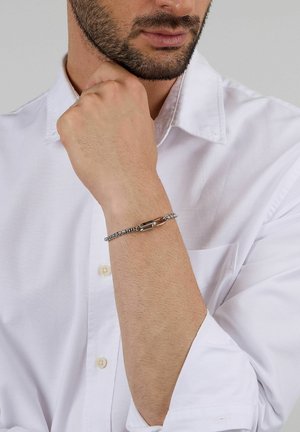Bracciale a catena d'argento con chiusura a moschettone, caratterizzato da maglie fini e finitura lucida, indossato su un polso nudo con una camicia bianca.
