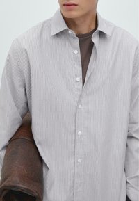 Gestreepte button-up shirt in lichtgrijs met witte accenten. Heeft een klassieke kraag, lange mouwen en een losse pasvorm. Bruin leren jasje over de arm gehangen.