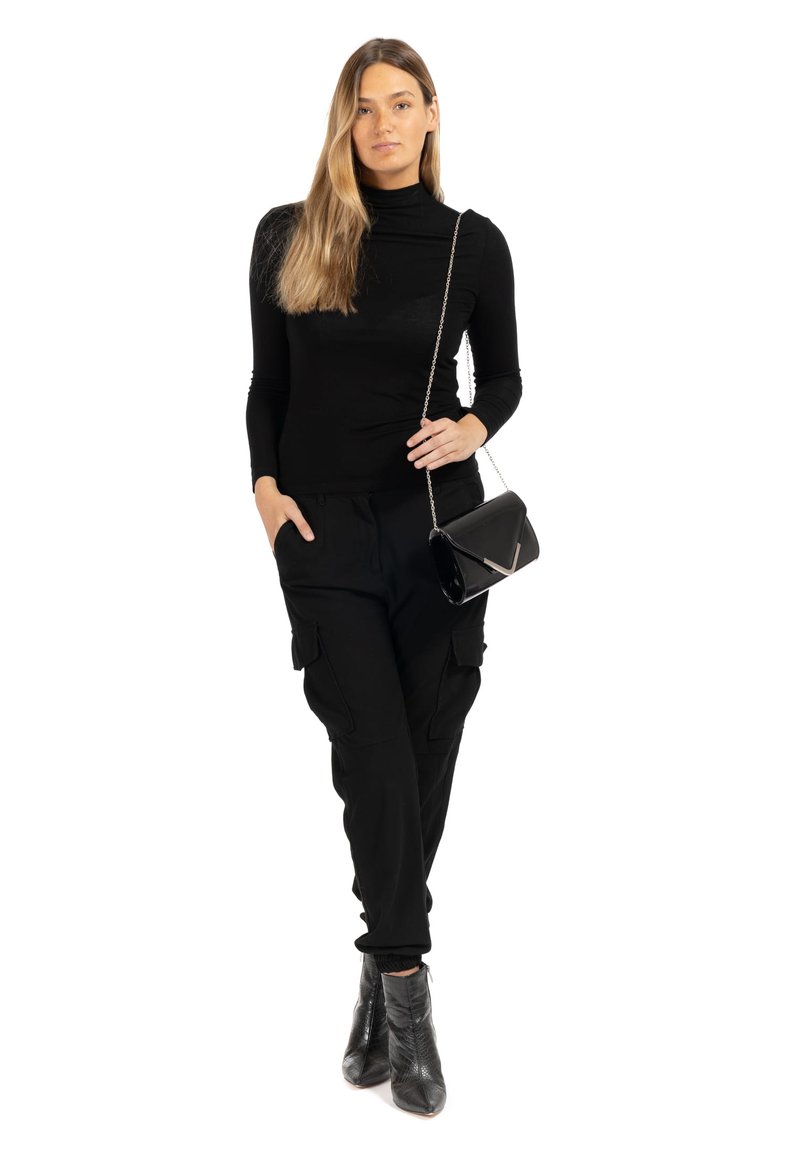 Femme aux longs cheveux blonds portant un col roulé noir, un pantalon cargo noir, des bottines noires et portant un petit sac noir avec une bandoulière en chaîne.