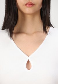 Top bianco con coste, dotato di scollatura a V profonda e dettaglio a occhiello al centro. Tessuto liscio e design aderente.