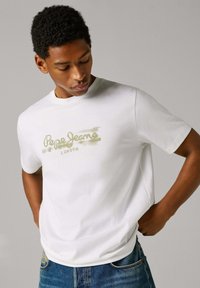 Λευκό βαμβακερό μπλουζάκι με στρογγυλή λαιμόκοψη, με πράσινη εκτύπωση σαν γκράφιτι "Pepe Jeans London" στο στήθος. Συνδυάζεται με μπλε τζιν παντελόνι.