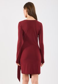 Vestido bordeaux de manga longa com um design ajustado, apresentando uma fenda lateral e um tecido liso e elástico. Vista traseira minimalista e elegante.