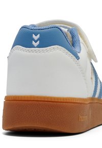 Biały sneaker z niebieską zakładką na pięcie, dwoma białymi szewronami, gumową podeszwą, widocznym przeszyciem i częściowo otwartym paskiem na rzep.