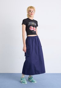 Black cropped T-shirt with a cherry graphic, paired with a long navy skirt featuring side pockets. Worn with light blue sneakers.