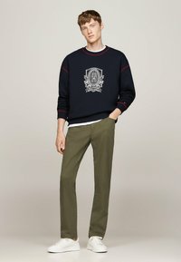 Felpa navy con cuciture rosse e grafica del logo, abbinata a pantaloni verdi e sneakers bianche, presenta una vestibilità rilassata e scollo rotondo.