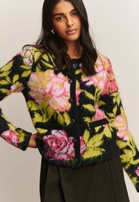 Femme portant un cardigan noir avec des motifs floraux roses et verts, des boutons noirs et des poches, associé à une jupe sombre.