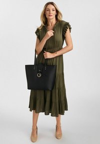 Czarna torba typu tote o gładkim wykończeniu, z dwoma długimi uchwytami, paskiem i okrąłym detalem w hardware. Noszona z oliwkową, warstwową sukienką.