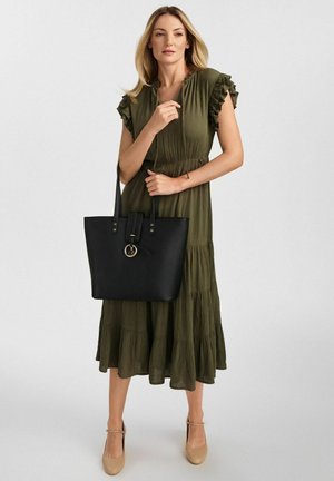 Czarna torba typu tote o gładkim wykończeniu, z dwoma długimi uchwytami, paskiem i okrąłym detalem w hardware. Noszona z oliwkową, warstwową sukienką.
