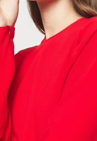 Blusa roja de tejido suave, con escote redondo y mangas largas que presentan un sutil detalle de costura. La textura parece suave y ligera.
