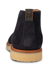 Botas de tobillo para hombre en ante azul marino con suela de goma color marrón, costuras azules y una trabilla en el empeine con una etiqueta marrón en la parte trasera.