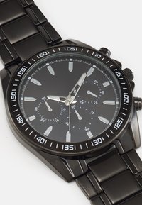 Montre chronographe noire avec un bracelet en métal noir, un cadran noir, des accents argentés et des index horaires luminescents. Équipée d'un affichage de la date et d'un tachymètre.