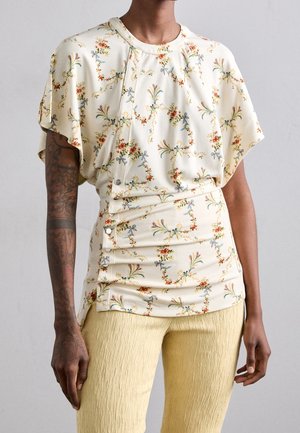 Persona con blusa beige de manga corta con flores y botones, y pantalones texturizados de color amarillo pálido, de pie con los brazos relajados a los lados.