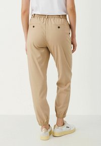 Khaki joggerbyxor med elastisk midja, två bakfickor med knappar och elastiska mudd, i kombination med vita sneakers och en T-shirt.