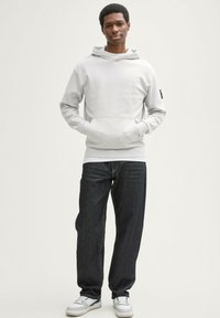 Felpa grigia chiara con tasca a marsupio, design allungato e polsini a costine, abbinata a jeans blu scuro e sneakers bianche.