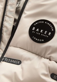 Puhdistava takki vaaleassa beige-kankaassa, jossa on pyöreä musta logopatch valkoisella tekstillä. Sisältää vetoketjusuljin ja kontrastiväriset ompeluyksityiskohdat.