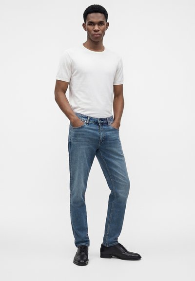 Un homme se tient debout, les mains dans les poches, portant une chemise blanche à manches courtes, un jean bleu et des chaussures habillées noires sur un fond clair uni.