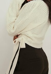 Strickpullover in Creme mit einem Bindeband in der Taille, getragen über einer schwarzen Hose. Die Textur ist locker mit einem weiten Ausschnitt und langen Ärmeln.