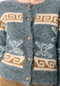 Frau trägt eine flauschige graue Strickjacke mit beigen geometrischen und Sternmustern, die vorne zugeknöpft ist, kombiniert mit braunen Cordhosen.