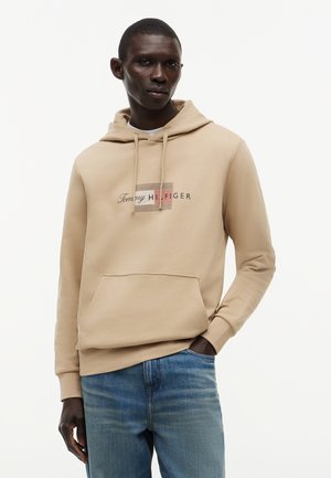 Άνδρας που φοράει μπεζ φούτερ Tommy Hilfiger και μπλε τζιν, στέκεται με το ένα χέρι στην τσέπη μπροστά σε απλό λευκό φόντο.