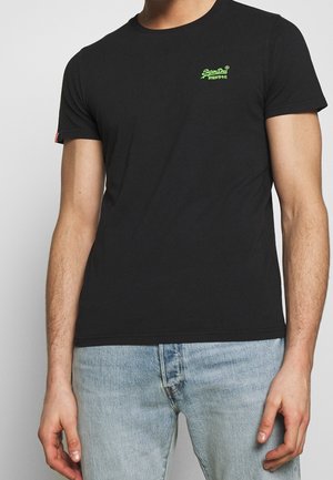 Mężczyzna w czarnej koszulce z krótkim rękawem z małym zielonym logo "SuperDry" i jasnoniebieskich dżinsach, stojący na jednolitym szarym tle.