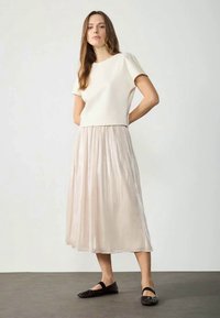 Cream short-sleeve top paired with a long, pleated, light beige skirt and black flat shoes. Smooth textures and simple design.