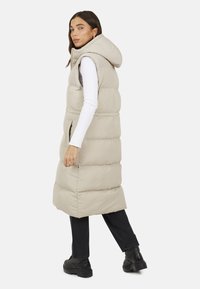 Gilet imbottito beige con cappuccio, design trapuntato, tasche laterali e silhouette lunga. Indossato sopra una maglietta bianca a maniche lunghe con pantaloni neri.
