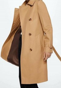 Beżowy płaszcz trencher o podwójnym zapięciu, z dużymi guzikami, bocznymi kieszeniami i paskiem w talii. Gładki materiał o średniej długości.