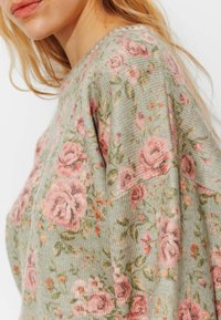 Pull fleuri avec un fond vert orné de roses roses et de feuilles, fabriqué en tissu texturé, avec un col rond et des manches larges.