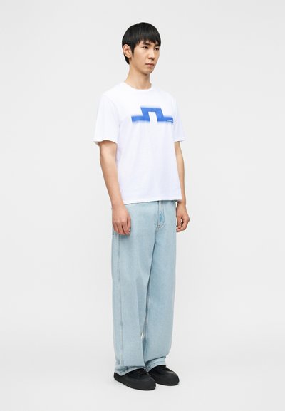 Λευκό βαμβακερό μπλουζάκι με μπλε γραφικό σχέδιο, συνδυασμένο με ανοιχτό μπλε τζιν παντελόνι με φαρδιές μπατζάκια και μαύρα παπούτσια με επίπεδη σόλα.