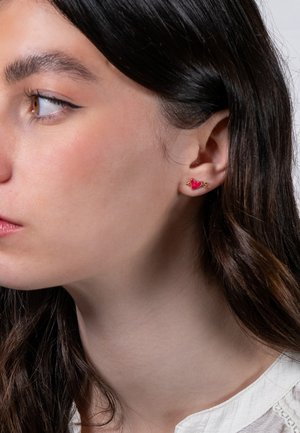 Orecchini d'oro con un piccolo design a cuore rosa brillante e delicate decorazioni, indossati all'orecchio di una persona. Lo sfondo è neutro.