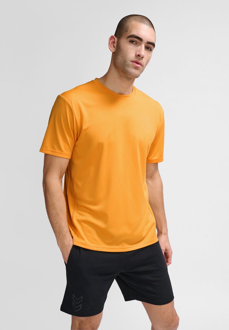 Orange atletisk t-shirt med korta ärmar och rund halsringning, med en diskret logotyp på vänster sida. Bärs med svarta shorts.