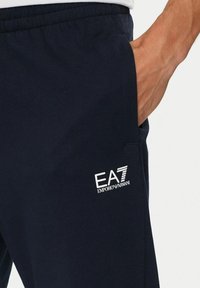Mano in tasca che indossa pantaloni della tuta Emporio Armani EA7 di un blu navy scuro con cintura elastica su sfondo bianco.