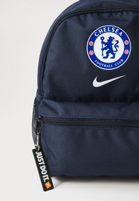 Navy blå ryggsekk laget av slitesterkt stoff med Chelsea Football Club-logo, Nike-merke og et "Just Do It"-tag. Har en glidelåslomme.