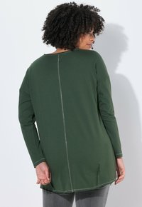 Haut vert à manches longues en tissu doux avec une coupe décontractée, présentant une couture centrale au dos et des coutures contrastées le long des bords.