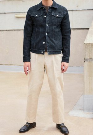 Person wearing a dark denim jacket, brown sweater, cream-colored trousers, and black loafers standing on a beige concrete floor.