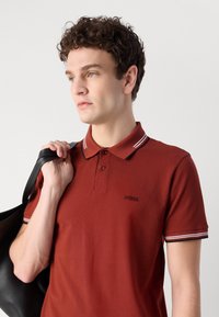 Červené polo tričko s trojici gombíkov. Má biele a čierne pruhované akcenty na golieri a rukávoch. Logo na hrudi.
