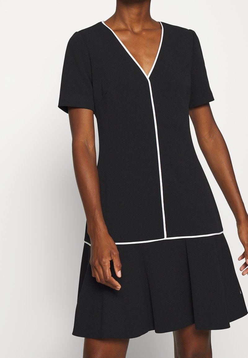 Femme portant une robe noire à manches courtes avec un passepoil blanc le long du col en V, de la couture frontale, de la taille et de l'ourlet.
