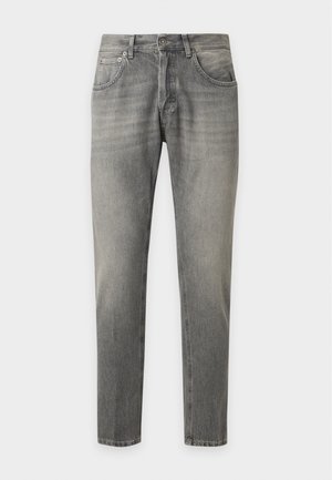 Jeune homme debout, les mains dans les poches, portant une chemise noire sur un t-shirt blanc, un jean gris et des baskets noires sur un fond blanc uni.