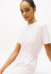 Roze katoenen jurk met logo, korte mouwen, elastische taille en een ontspannen pasvorm. Glad textuur tegen een witte achtergrond.