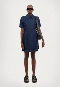 Czarna sukienka z denimu z krótkimi rękawami, spiczastym kołnierzem i widocznymi szwami. Modelka nosi czarne okulary przeciwsłoneczne i czarną torbę na ramię.