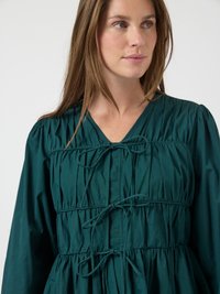 Donna che indossa un abito lungo a maniche lunghe di colore verde scuro con tessuto arricciato e tre fiocchi legati sulla parte anteriore, mentre guarda di lato su uno sfondo bianco.