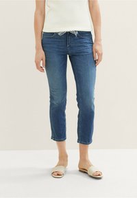 Mujer con jeans azules recortados y cintura con cordón, camiseta sin mangas de punto blanca y sandalias beige, de pie sobre un suelo blanco.