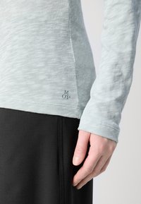 Šviesiai mėlynas ilgomis rankovėmis viršus iš tekstūruotos medžiagos, su kreivu apačios kraštu ir logotipo detale. Suderintas su juodomis kelnėmis, matoma dalis rankovės.