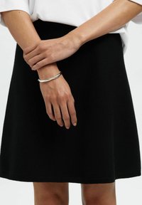 Schwarzer Strickrock mit einem ausgestellten Design, weicher Textur und elastischem Bund. Ein silbernes Armband schmückt eine Hand, die auf dem Rock ruht.