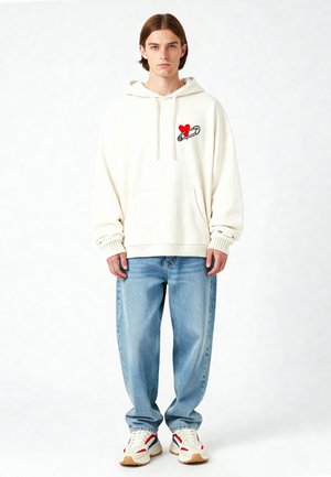 Crème hoodie met een grafisch ontwerp van een rode hart en een veiligheidsspeld, gecombineerd met lichtblauwe loszittende jeans en beige sneakers met rode en blauwe accenten.